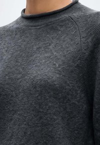 Gros plan d'une personne portant un pull en tricot gris foncé avec un col rond côtelé et des détails de points texturés près de l'épaule.