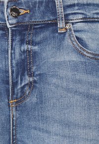 Primer plano de bolsillo de jeans de mezclilla azul con costuras naranjas, botón metálico y presilla para cinturón.