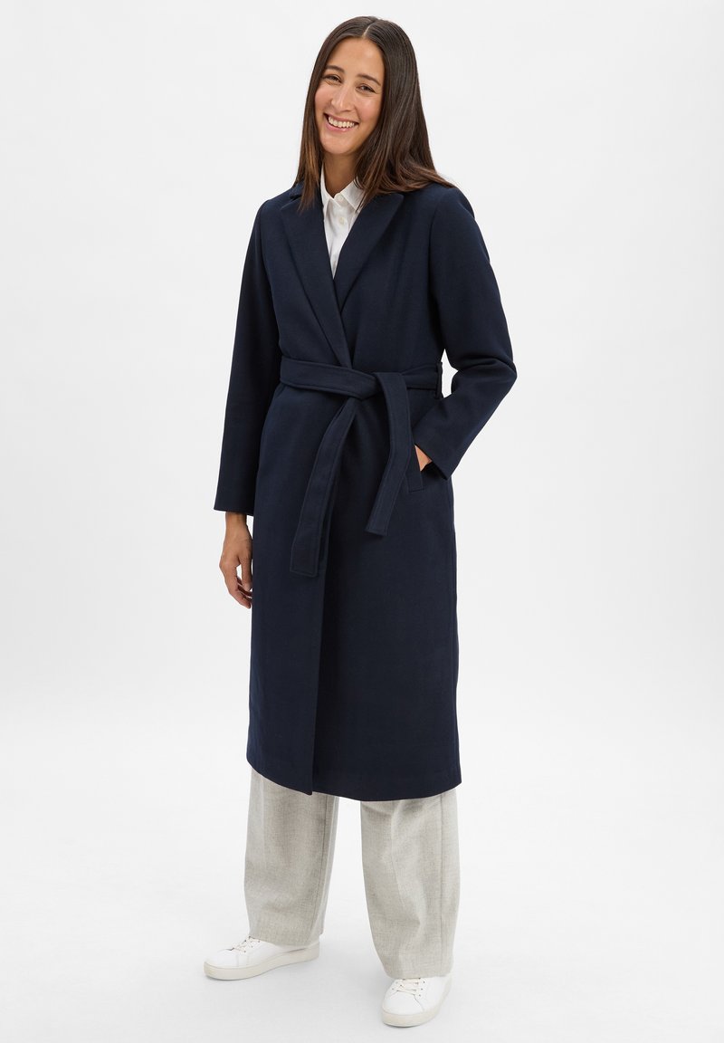 Marineblauer langer Mantel mit Wickeldesign, aus einem weichen Material gefertigt. Mit einem Gürtel, seitlichen Taschen und langen Ärmeln. Getragen über hellen Hosen.