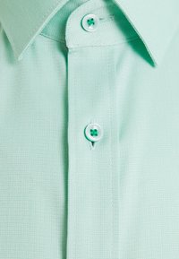 Gros plan d'une chemise texturée vert clair avec un col pointu et deux boutons visibles, un fixé près du col et un en dessous.