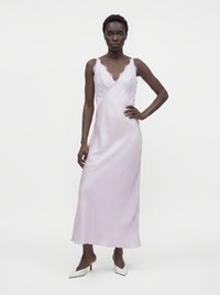 Femme portant une robe nuisette en satin violet clair avec des détails en dentelle, debout avec une main sur la hanche et des talons blancs pointus aux pieds.