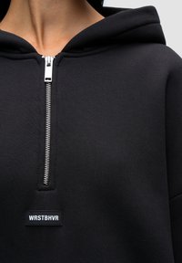 Czarna bluza z kapturem, z przednim zamkiem błyskawicznym, prążkowaną fakturą i prostokątną naszywką z etykietą "WRSTBHVR" wszytą na przodzie.