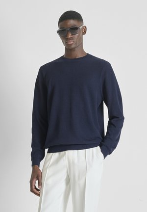 Uomo che indossa un maglione a maniche lunghe blu scuro, pantaloni bianchi plissettati e grandi occhiali da sole neri, in piedi davanti a uno sfondo neutro.