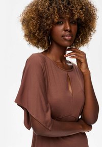 Vrouw met krullend haar, gekleed in grote oorbellen en een bruine top met fladdermouwen, met één hand op haar kin en een zelfverzekerde uitstraling.