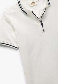 Hvid polo-shirt lavet af tekstureret bomuld med lynlås. Har et kontrasterende marineblå stribe på kraven og ærmeenderne.