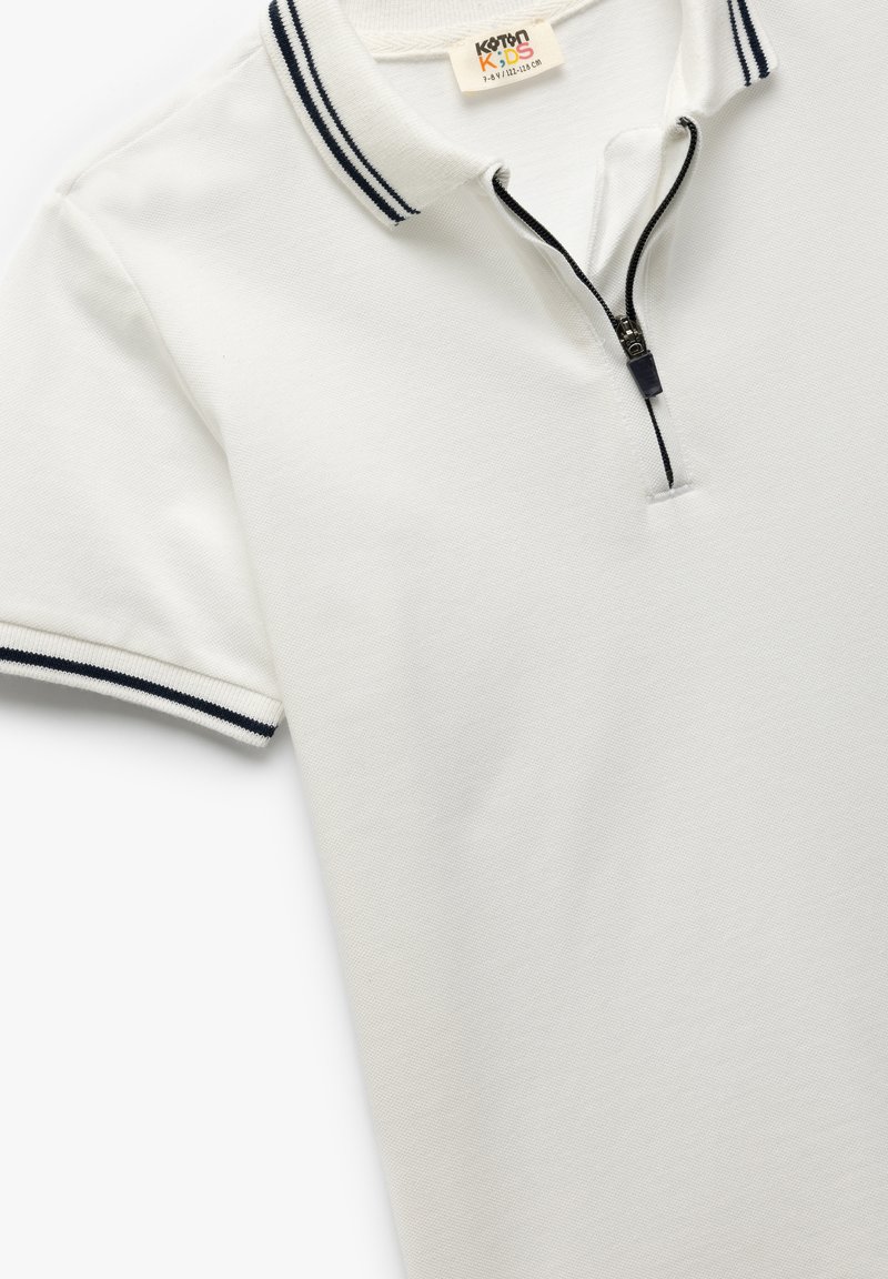 Hvid polo-shirt lavet af tekstureret bomuld med lynlås. Har et kontrasterende marineblå stribe på kraven og ærmeenderne.