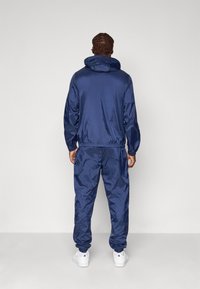 Marineblauwe trainingsoutfit van glanzend, soepel materiaal. Voorzien van een capuchon, elastische manchetten en tailleband, gecombineerd met witte sneakers.