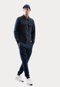 Giacca navale con zip e due tasche sul petto, abbinata a pantaloni tapered coordinati e sneakers bianche. Il tessuto appare liscio e morbido.