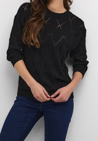 Schwarzer Strickpullover mit einem Diamantenmuster und offenen Maschen, Rundhalsausschnitt sowie gerippten Bündchen und Saum, kombiniert mit dunklen Jeans.