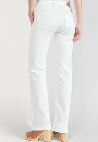 Jean en denim blanc évasé avec une taille haute, comportant deux poches arrière et un motif de couture. Porté avec des sabots en bois de couleur claire.