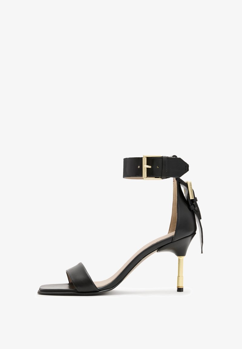 Black leather heeled sandal with a square toe, ankle strap featuring a gold buckle, and a slender metallic heel. Smooth texture, minimalist design.