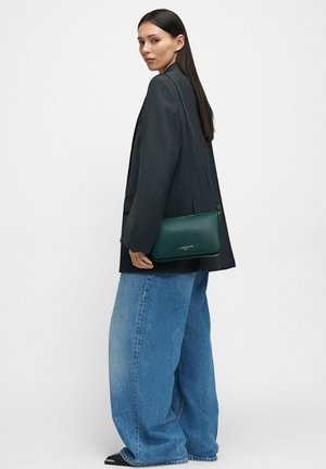 Borsa a tracolla in pelle verde con chiusura a zip, forma rettangolare e logo discreto in dettaglio metallico, indossata sopra un blazer nero e jeans larghi.