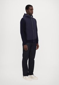 Marinblå hooded jacka med dragkedja fram, ribbade ärmslut och nederkant, i kombination med mörka jeans och vita sneakers. Slät tygtextur, avslappnad design.