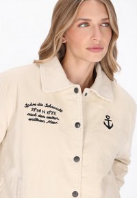 Beige corduroy jakke med krave, med sorte knapper, broderet tekst og et ankeremblem fortil. Glat tekstur.