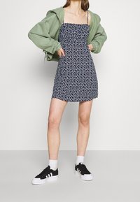 Námořnické šaty s bílými puntíky, nařasený výstřih, tenké ramínka, spárované s oversized zelenou bundou a černými teniskami s bílými pruhy.