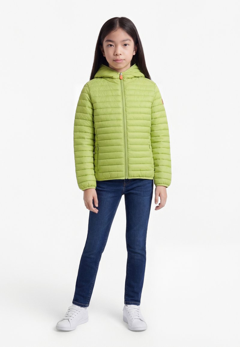 Lime zöld kapucnis pufidzsekiben, sötétkék farmerben és fehér tornacipőben álló lány egyszínű fehér háttér előtt.