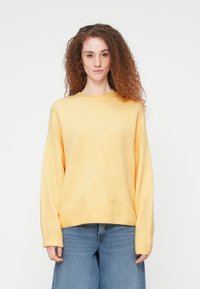Jeune femme aux longs cheveux bouclés portant un pull jaune ample et un jean large bleu, se tenant devant un fond blanc uni.