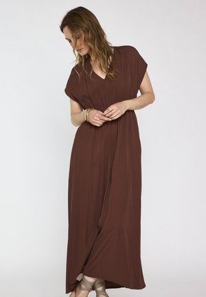 Femme aux cheveux ondulés portant une robe maxi marron ample et des chaussures à lacets à motifs, regardant vers le bas avec les mains jointes devant.