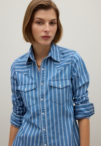 Giovane donna con capelli corti castani che indossa una camicia a righe verticali blu e bianche con le maniche arrotolate su uno sfondo semplice.
