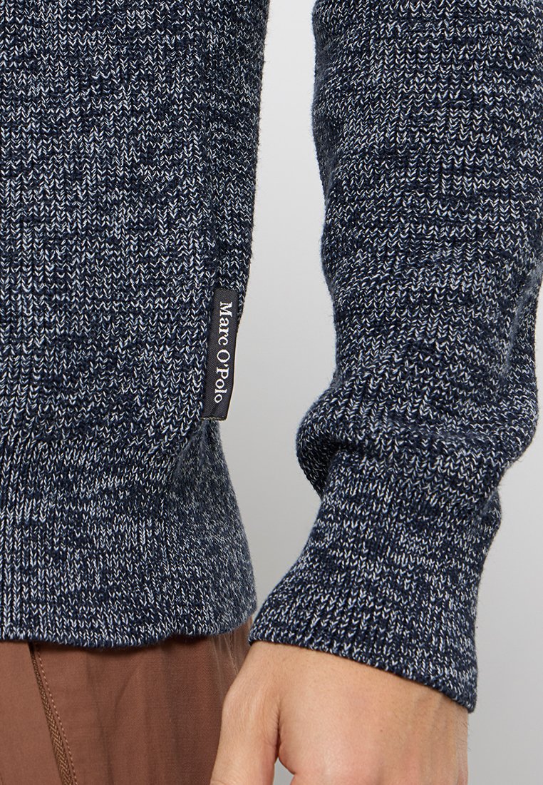 Nahaufnahme eines Arms einer Person, die einen blau-weißen Strickpullover mit einem Marc O'Polo-Label trägt, kombiniert mit braunen Hosen.