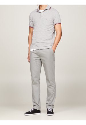 Homme portant un polo gris clair, un pantalon gris clair assorti, des baskets noires avec des lacets blancs, debout avec une main dans la poche.