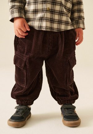 Kind draagt bruine corduroy broek met cargozakken, een geruit overhemd en zwarte sneakers met bruine zolen, staand op een lichte vloer.