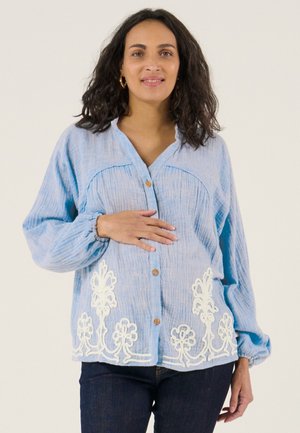 Vrouw met donker krullend haar draagt een lichtblauwe blouse met witte bloemembroderie, knoopsluiting aan de voorkant, en donkere spijkerbroek, hand rust op de buik.