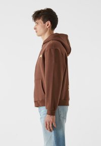 Ruda megztinė su kapuce, su kengūros kišene, raukšlėtomis rankovėmis ir apačia. Išskirtinis auksinis logotipas ant krūtinės, derinamas su šviesiai mėlynomis džinsinėmis kelnėmis.