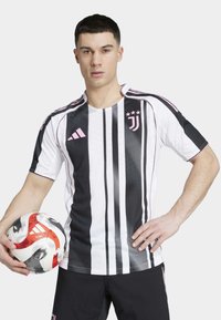 Maillot de soccer rayé noir et blanc avec des accents roses, à manches courtes et col rond, porté par un mannequin tenant un ballon de soccer.