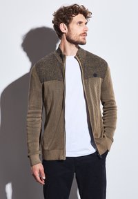 Zipped cardigan in earthy brown with textured upper panel. Features ribbed pattern and long sleeves. Paired with a white T-shirt underneath.