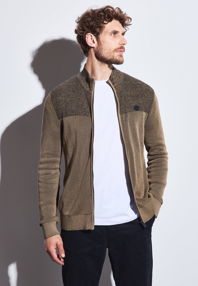 Zipped cardigan in earthy brown with textured upper panel. Features ribbed pattern and long sleeves. Paired with a white T-shirt underneath.