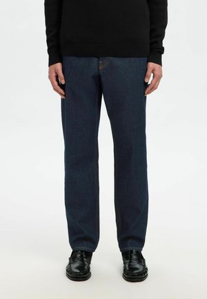 Personne portant un jean droit bleu foncé, des mocassins en cuir noirs et un pull noir à manches longues, debout devant un fond uni.