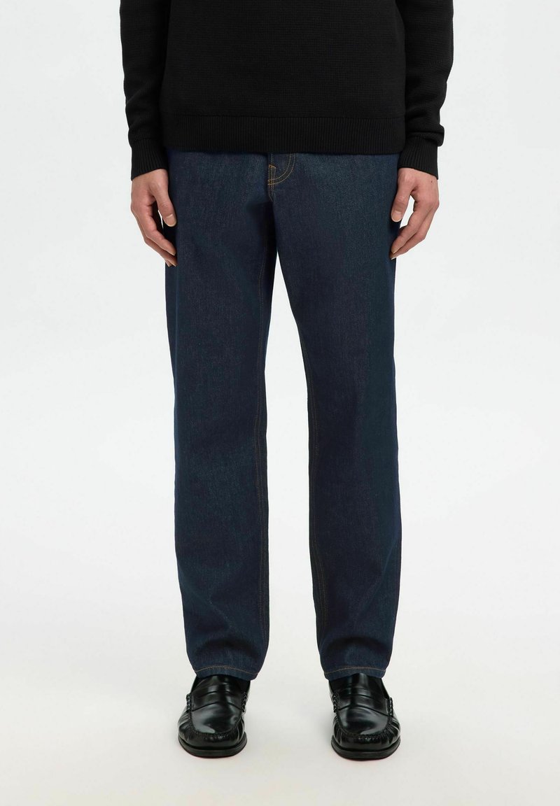 Personne portant un jean droit bleu foncé, des mocassins en cuir noirs et un pull noir à manches longues, debout devant un fond uni.