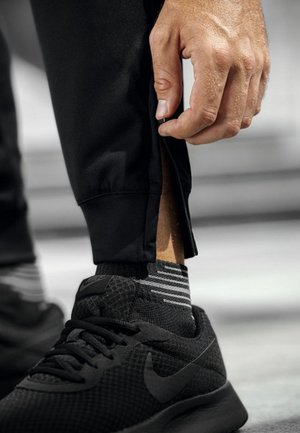 Schwarze sportliche Jogginghosen mit einem Reißverschluss am Bündchen, kombiniert mit schwarzen Sneakers und gestreiften grauen Socken. Die Jogginghosen haben eine glatte Textur.