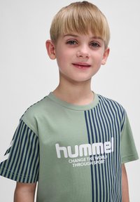 T-shirt en coton vert avec des rayures verticales bleu marine, un col rond et un texte blanc qui dit "hummel CHANGE THE WORLD THROUGH SPORT."