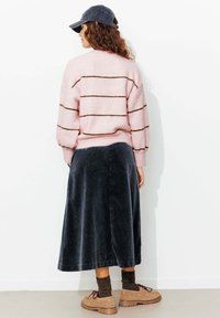 Pull rose clair en maille avec des rayures marron, assorti à une jupe en velours côtelé foncé. Le mannequin porte une casquette foncée et des chaussures beiges épaisses.