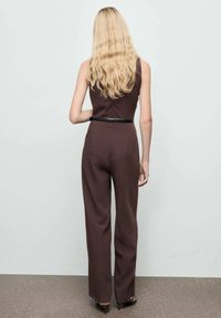 Bruine mouwloze jumpsuit met een smalle pasvorm en wijde broekspijpen, accentueert door een dunne zwarte riem, vanaf de achterkant getoond.