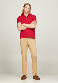 Polo rosso a maniche corte, pantaloni chinos beige e scarpe marroni. La maglietta presenta un piccolo logo, realizzata in tessuto morbido, con una vestibilità su misura.