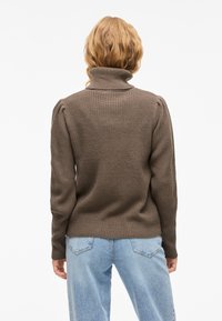 Pulover din tricotaje bej cu guler înalt, cu umeri bufanți, purtat cu blugi de un albastru deschis. Textură moale, design ajustat și mâneci de lungime medie.