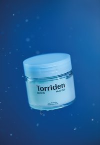 Torriden Dive In Multi Pad i et klart glasburk med et lyseblåt låg, prydet med sort tekst, svævende i vanddråber mod en blå baggrund.