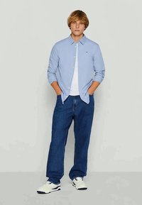 Camisa de botões azul clara sobre uma t-shirt branca, combinada com jeans azul escuro e sapatilhas brancas. Ajuste casual, mangas longas, colarinho padrão.