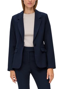 Mörkblå kavaj med slag, enknappsstängning, två framfickor och en skräddarsydd passform, i kombination med en ljusbeige topp.