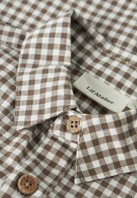 Hnědobílá kostkovaná košile s límečkem, dřevěnými knoflíky a štítkem s nápisem "Lil'Atelier". Viditelný detail texturované látky.