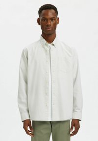 Ανοιχτό πράσινο ριγέ πουκάμισο με τσέπη, μακριά μανίκια και κλασικό γιακά, φορεμένο πάνω από ένα λευκό φανελάκι, σε συνδυασμό με ανοιχτόχρωμο παντελόνι.