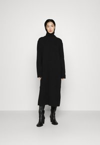 Schwarzes Strickkleid mit hohem Kragen, langen Ärmeln und Seitenschlitzen, kombiniert mit enganliegenden schwarzen Lederstiefeln bis zum Knie. Glatte Textur.