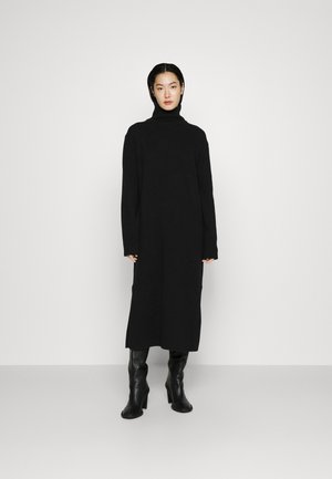 Schwarzes Strickkleid mit hohem Kragen, langen Ärmeln und Seitenschlitzen, kombiniert mit enganliegenden schwarzen Lederstiefeln bis zum Knie. Glatte Textur.
