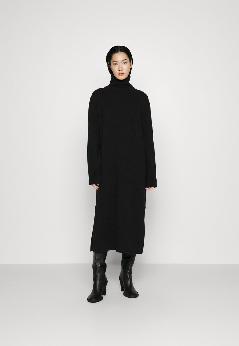 Schwarzes Strickkleid mit hohem Kragen, langen Ärmeln und Seitenschlitzen, kombiniert mit enganliegenden schwarzen Lederstiefeln bis zum Knie. Glatte Textur.