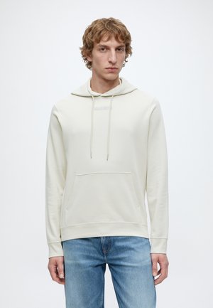 Young man with wavy blond hair wearing a light beige hoodie and blue jeans, standing against a plain white background.