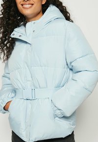 Lyseblå polstret jakke med hætte, bælte i taljen, elastiske ærmeafslutninger og glat stof. Har lynlås og trykknapper foran.