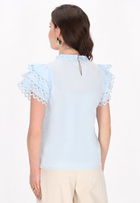 Blusa azzurra chiara con maniche a volant caratterizzata da ritagli circolari, colletto alto e design con apertura a chiave sul retro. Tessuto liscio e leggero.